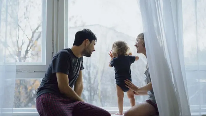 Happy family with young cute daughter sitting on windowsill playing and looking in window at home